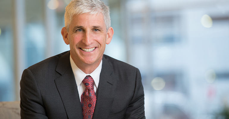white haired man wearing a suit and tie smiles in front of a blurred background.