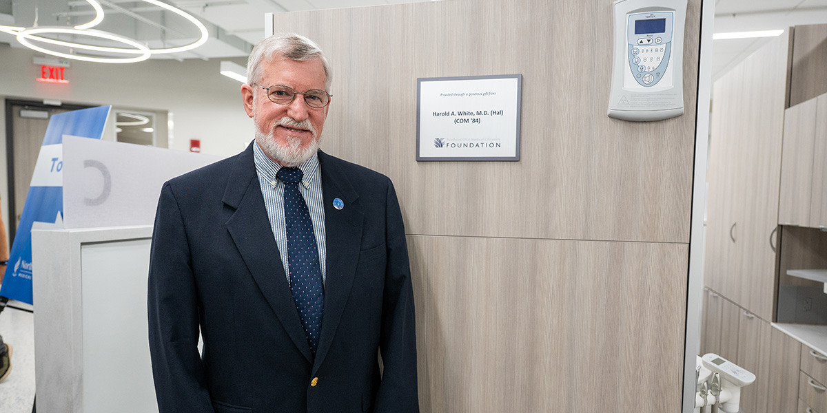 Man in suit and tie poses next to a plaque outside a dental operatory space.