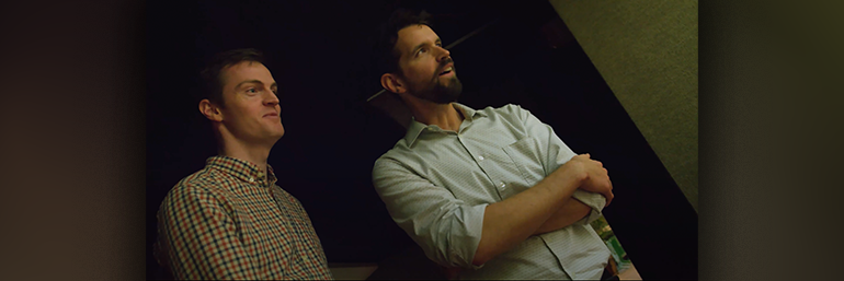 Man in plaid shirt and man in white shirt are watching something out of frame