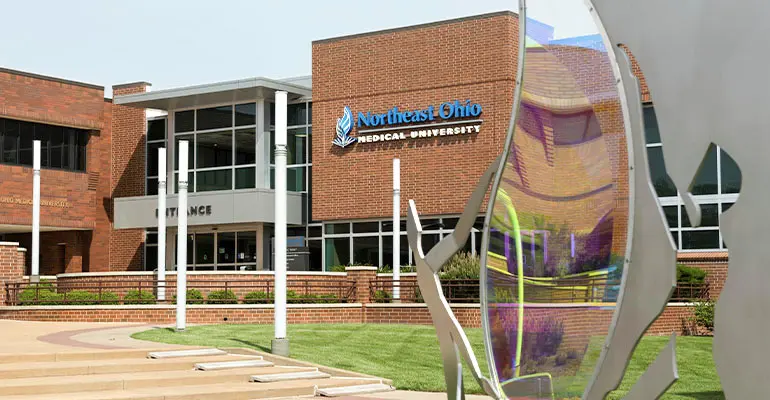 The entrance of Northeast Ohio Medical University, featuring a modern brick building with the university’s name and logo displayed, white columns at the entryway, green landscaping, and a reflective outdoor sculpture in the foreground.