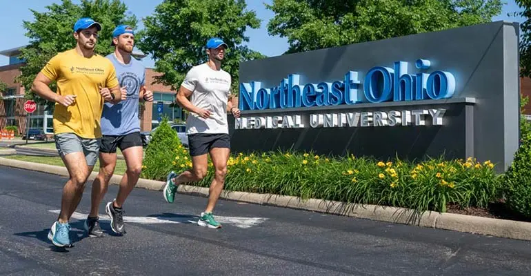 Three medical students who are training for an ultramarathon run by a NEOMED sign on campus.