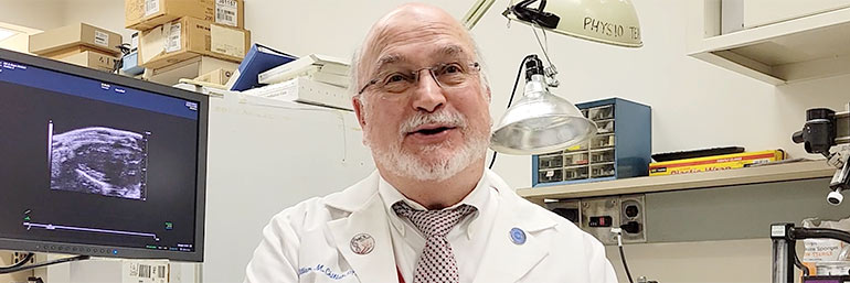 A NEOMED researcher in a white coat in a lab with a screen over his shoulder.