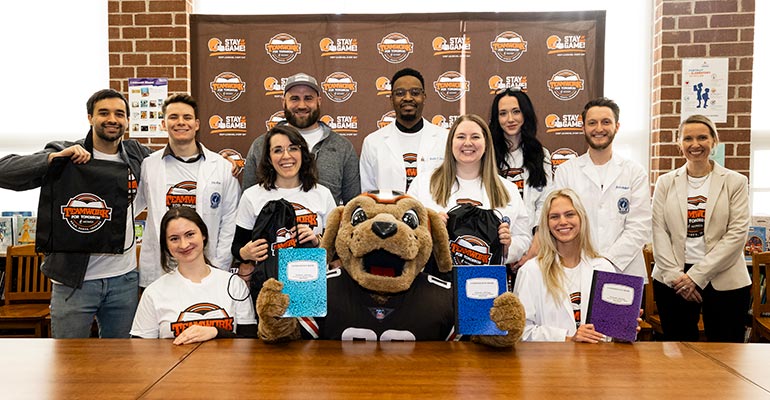 NEOMED College of Pharmacy students gather with the Browns mascot for a photo around a table with books.