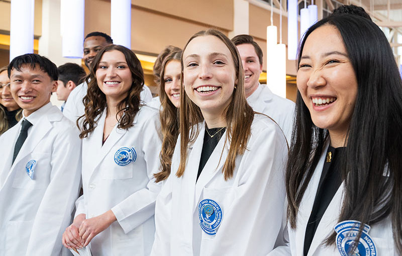 A group of smiling anesthesia assistant students stand in a group, wearing their white coats for the first time.