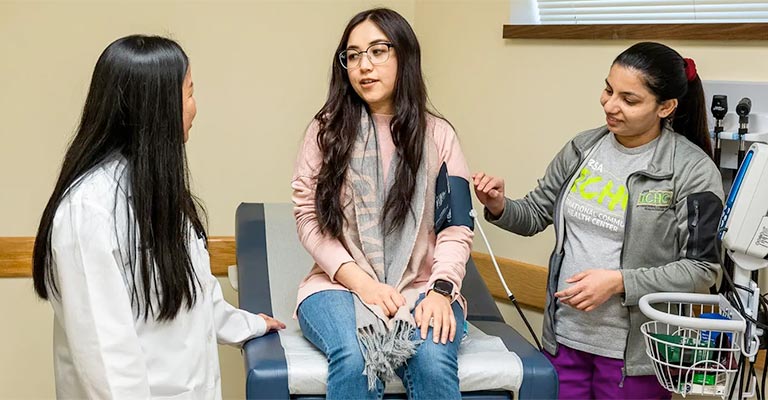 A doctor in a white coat chats with a patient and her mother in an examination room.