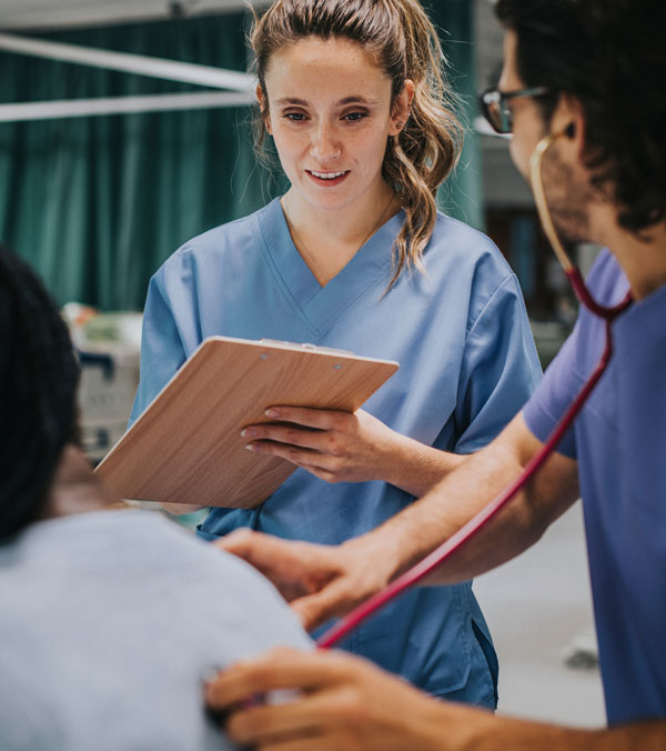 A mental health professional in blue scrubs holds a clipboard while collaborating with colleagues during a patient examination.