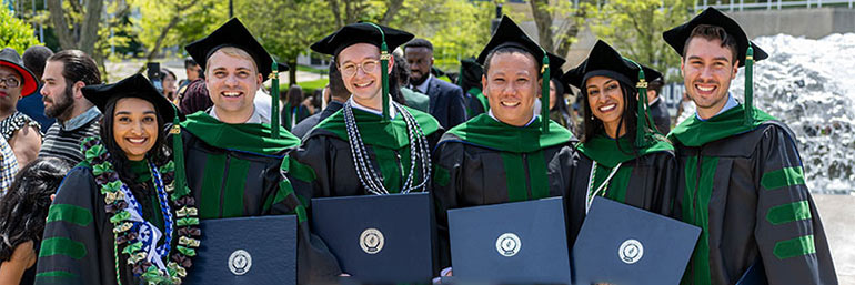 Six NEOMED graduates stand in their commencement robes outside of E.J. Thomas Performing Arts Hall.