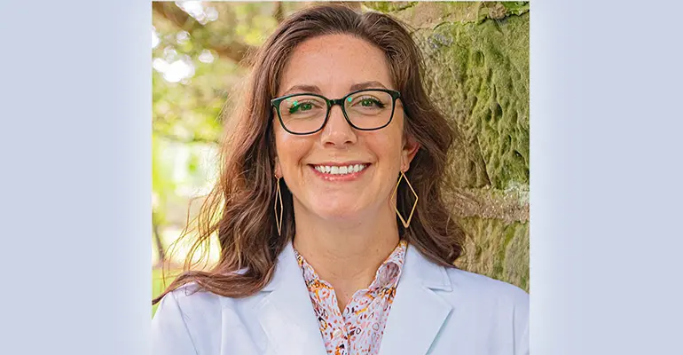 A woman wearing glasses and a white coat smiles outdoors beside a moss-covered stone wall, with sunlight filtering through trees in the background.