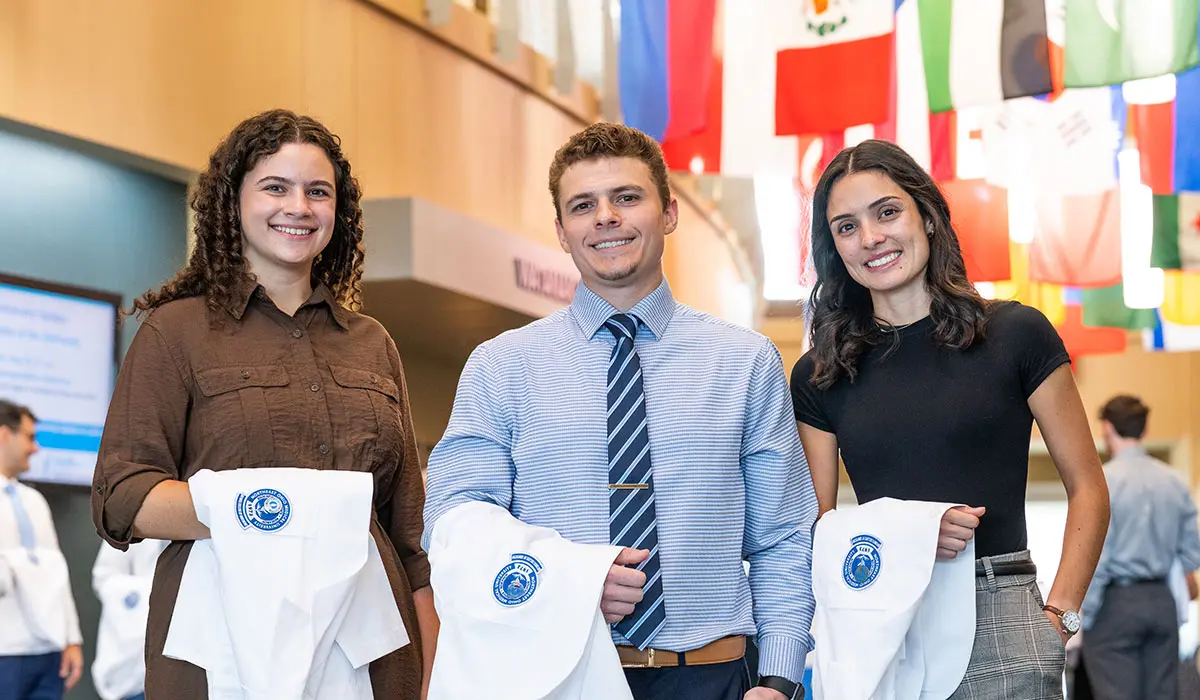 Three dentistry students stand together indoors holding white coats embroidered with the NEOMED College of Dentistry logo, smiling as international flags hang above them.
