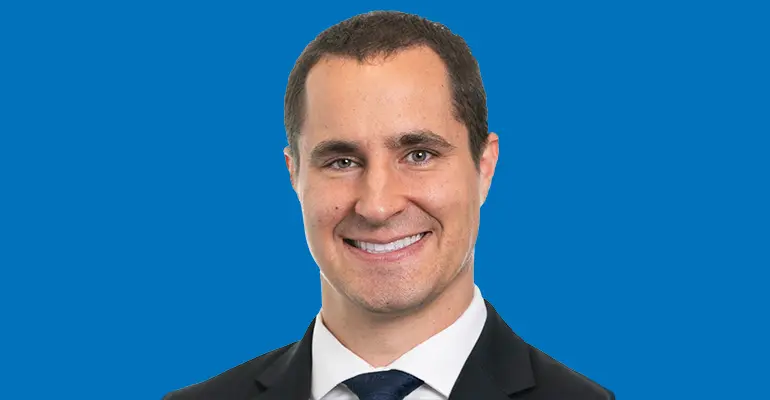 Professional headshot of a smiling man in a dark suit, white shirt and navy tie, posed against a solid blue background.