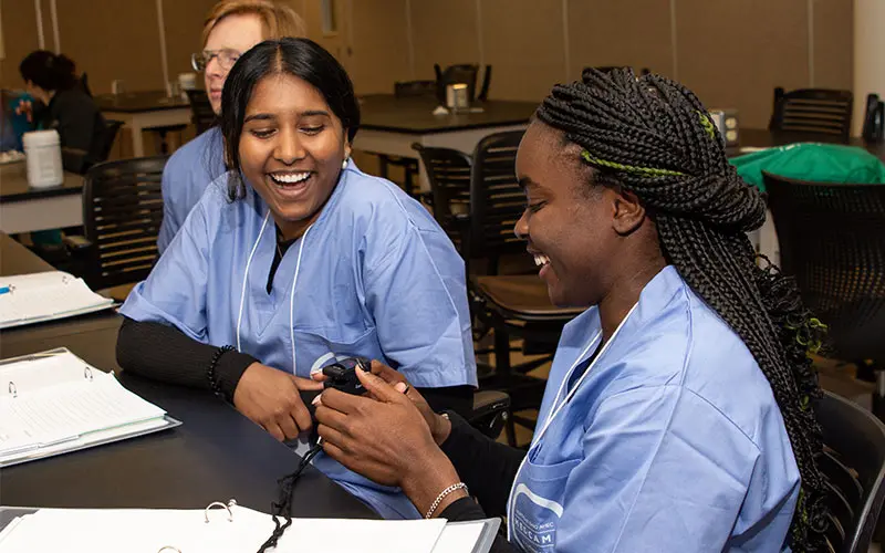 Two participants in NEOMED’s Health Careers Bootcamp, wearing blue scrubs, sit at a classroom table smiling and laughing as they examine a handheld medical device during a learning activity.