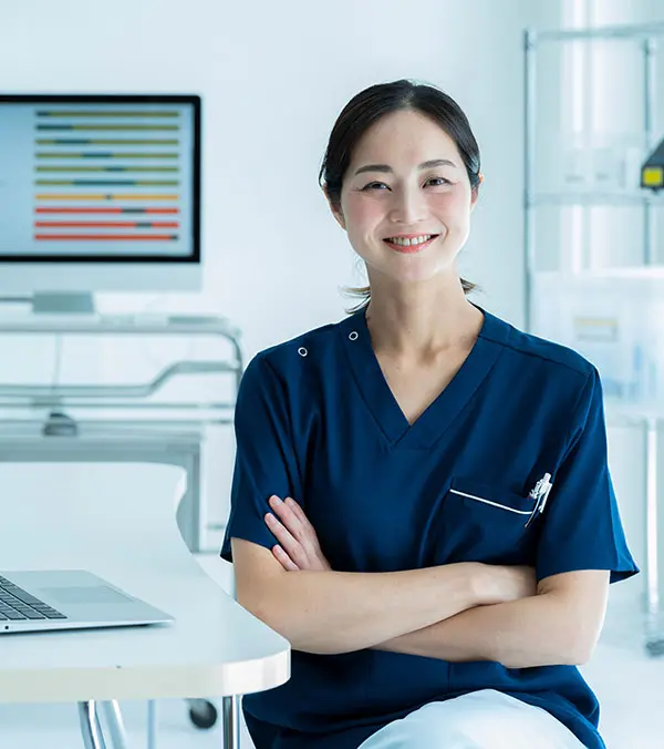 A smiling healthcare researcher in navy scrubs sitting with arms crossed in a bright medical office.
