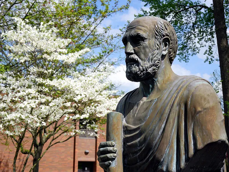 Bronze statue of Hippocrates holding a scroll, set outdoors in front of a brick building and blooming white dogwood trees under a partly cloudy sky.