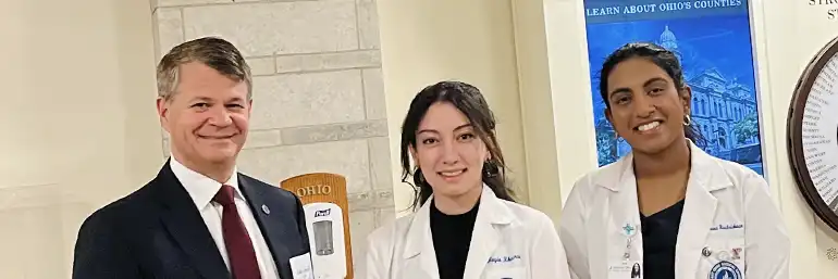 Two NEOMED medicine students in white coats stand with NEOMED's president in Columbus.