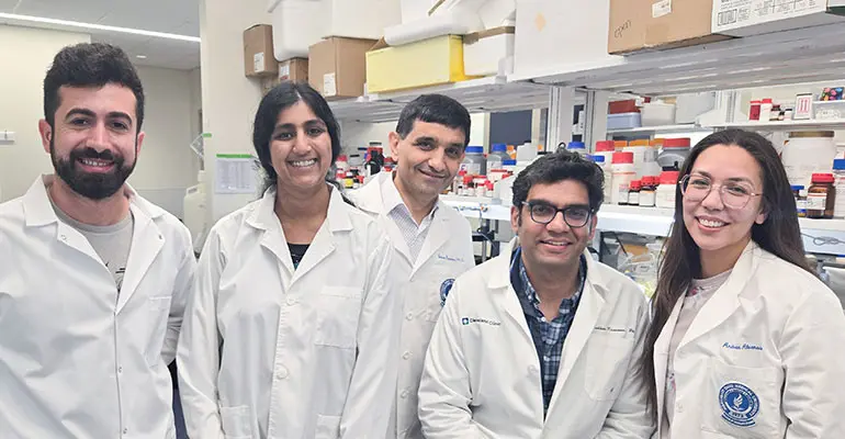 A group of five researchers wearing white lab coats smile while standing together in a laboratory filled with scientific equipment, shelves of bottles, and research supplies.