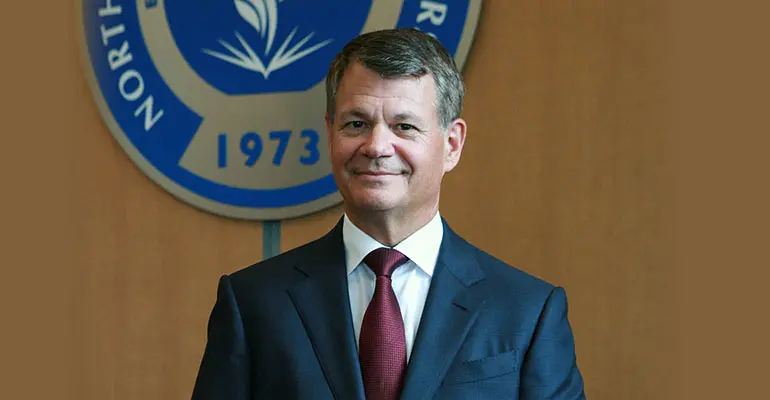 A man in a dark suit with a red tie is standing in front of a wall displaying part of the University's crest.