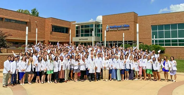 The College of Medicine's Class of 2028 stand in white coats in front of the main NEOMED entrance.