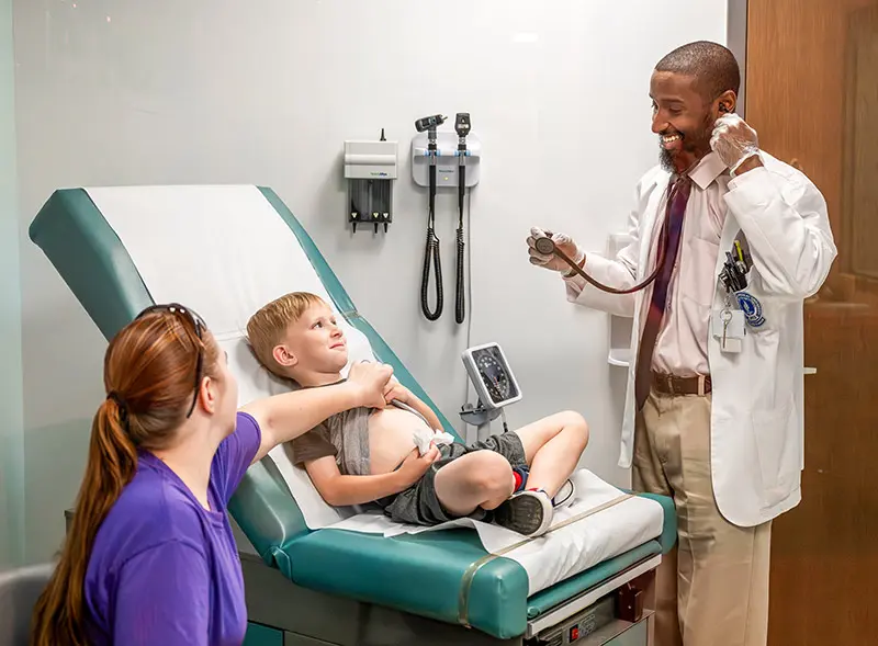 A student doctor talks with a mother and a young child in an examination room.