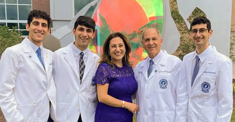 A group of five people stands smiling outdoors in front of a colorful sculpture, with three young men and one older man wearing white medical coats and a woman in a purple dress at the center.