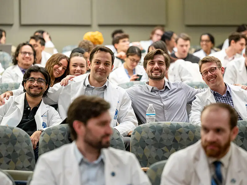 A group of medical students, many wearing white coats, sit together in a lecture hall smiling and talking before class, creating a collegial atmosphere.