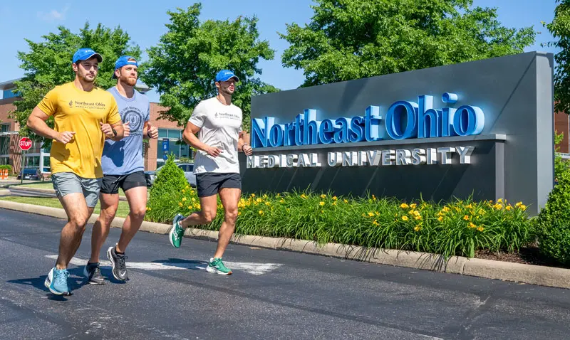 Three runners jog past the NEOMED sign at the University's main entrance on a warm, sunny day.