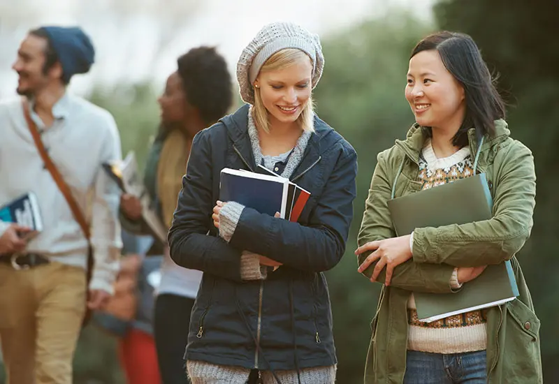 Students in light coats walk on campus, talking.