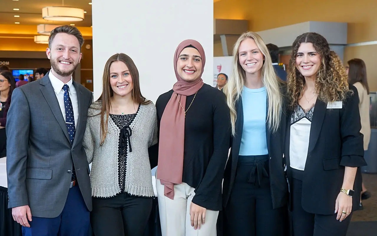 A group of five pharmacy students stand together smiling in a brightly lit lobby during an on-campus career fair, dressed in professional attire with other attendees visible in the background.