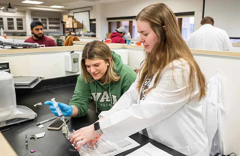 Two students work together in a laboratory, one wearing a white coat and the other in a green sweatshirt, as they prepare materials with syringes and lab equipment.