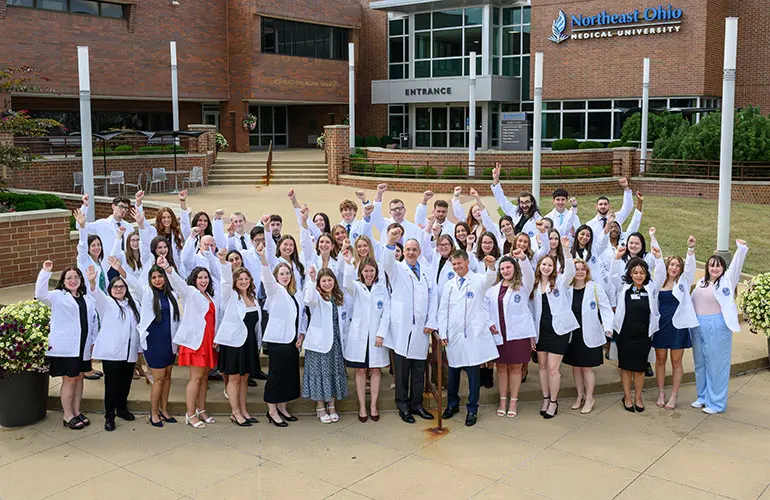 A large group of pharmacy students and faculty in white coats stand smiling with raised arms in front of the Northeast Ohio Medical University entrance.