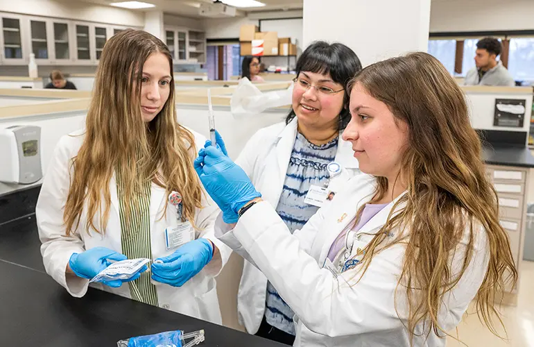 Three people in lab coats stand together in a pharmacy lab, with one holding up a syringe while the others observe and discuss.