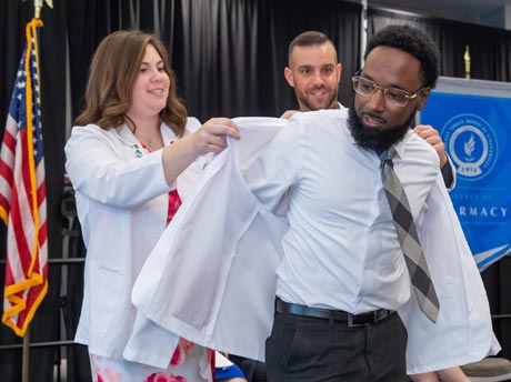 Two pharmacy students help new pharmacy student put on his white coat for the first time.