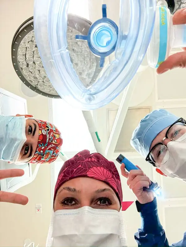 Three NEOMED Certified Anesthesiologist Assistant students in surgical masks and caps look down toward the camera while holding an anesthesia mask and medical tools in an operating room.