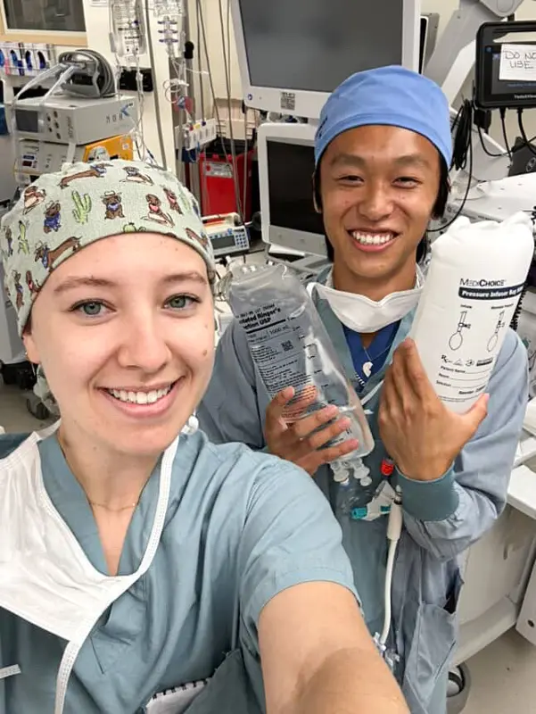 Two Certified Anesthesiologist Assistant (CAA) students in scrubs and surgical caps smile in a clinical setting while holding IV fluid bags, with hospital equipment visible behind them.