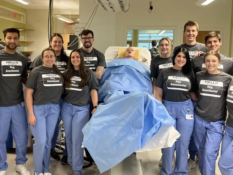  Group of certified anesthesia assistant (CAA) students in scrubs and matching “Hello, Hey Anesthesia!” T-shirts standing around a hospital bed with a medical mannequin, inside a clinical simulation lab.