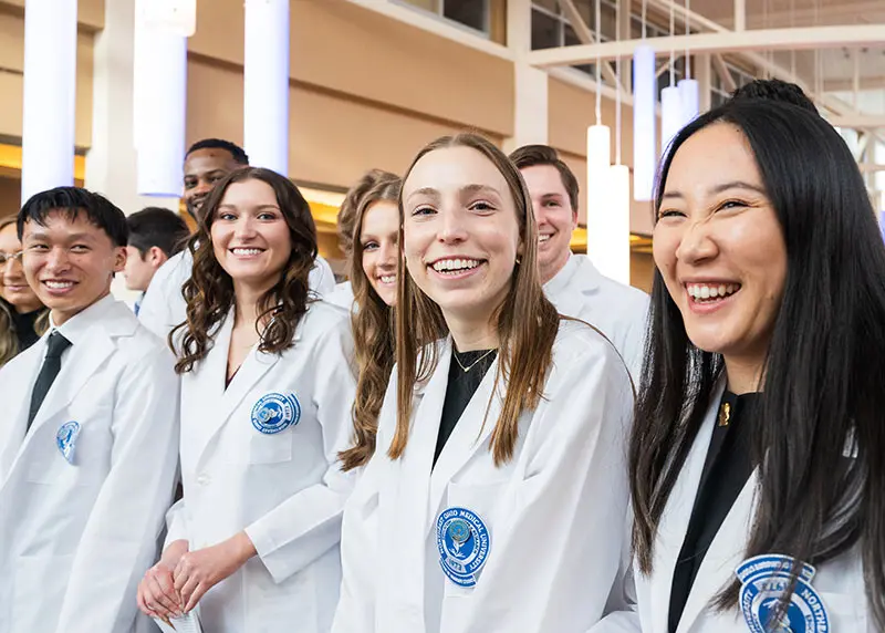 A group of students in white coats stand together indoors, smiling after the White Coat ceremony.