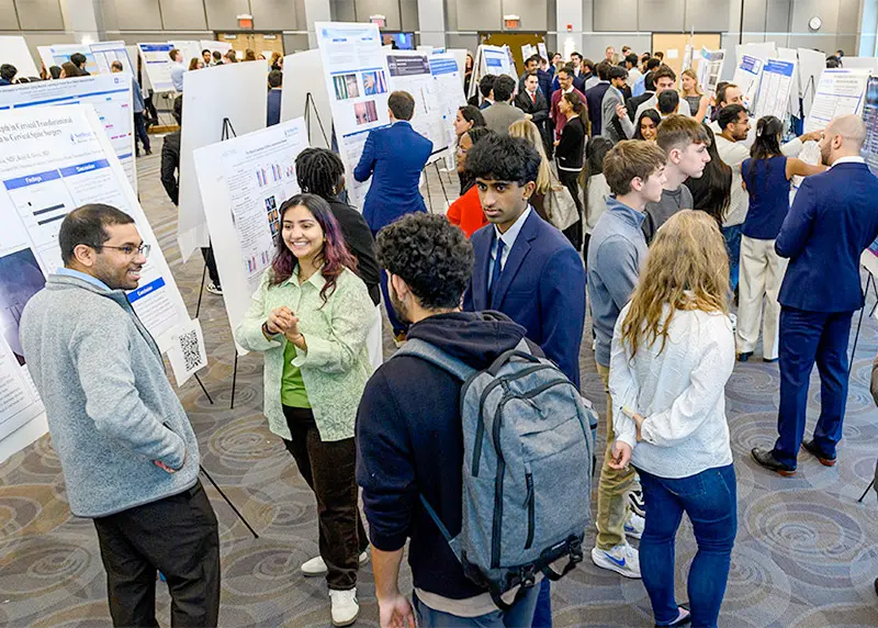 Students and faculty mingle and discuss research posters in a crowded presentation hall.