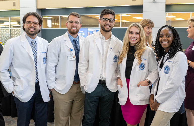 Five medical students in white coats chat during a campus event.