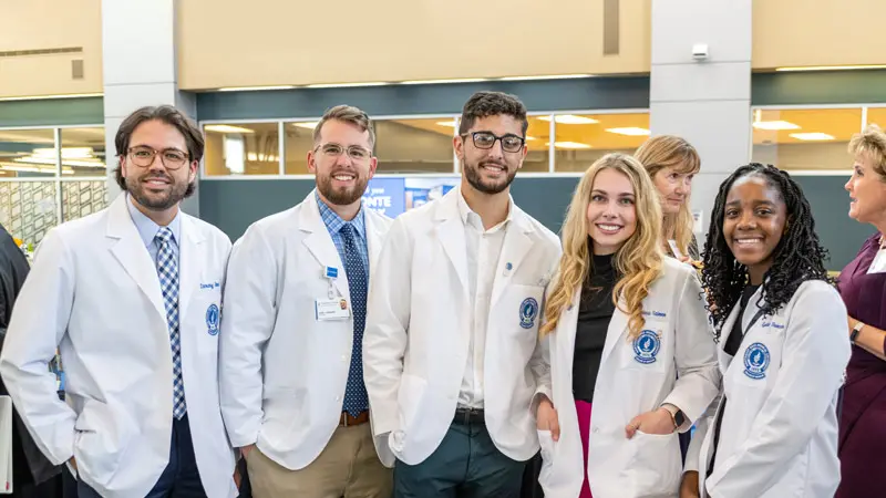 A group of NEOMED students wearing white coats stand together indoors, smiling during a campus event.