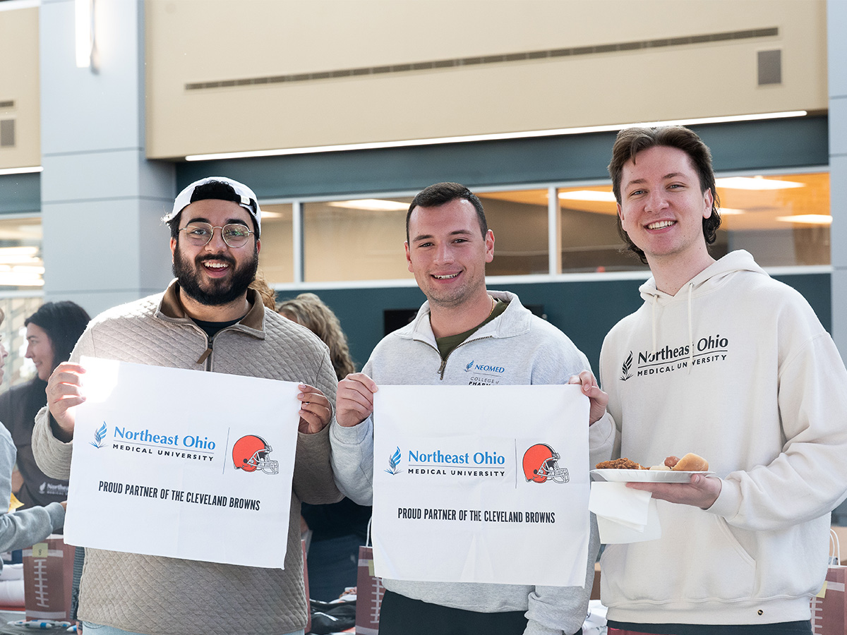 Three male NEOMED students smiling with two holding sign with co-branded logo and the last student holding a plate with a hotdog on it