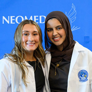 Two pharmacy students in white coats stand before a banner that reads NEOMED.