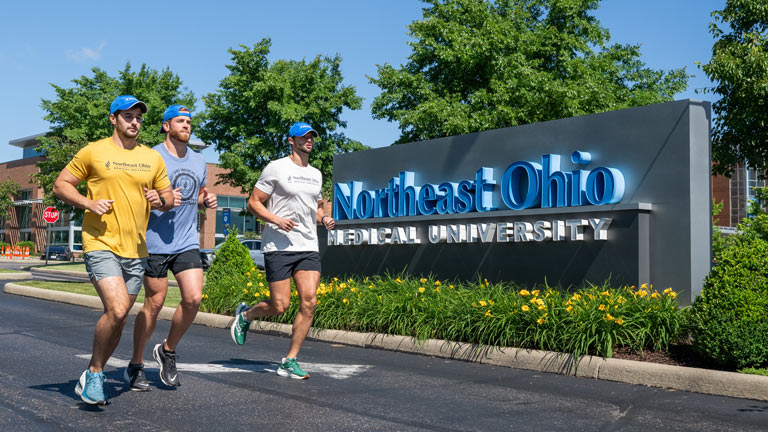 Three medical students in running gear jog past the NEOMED sign in front of the university.