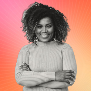 A smiling woman in a tan sweater stands with her arms crossed.